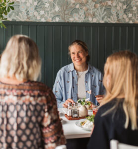 Foto. En kvinna i uppsatt hår sitter vid bordet och ler mot sina bordvänner.