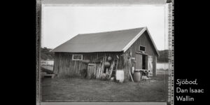 Svartvitt foto. Profilbild av en gammal sjöbod, havet i bakgrunden.