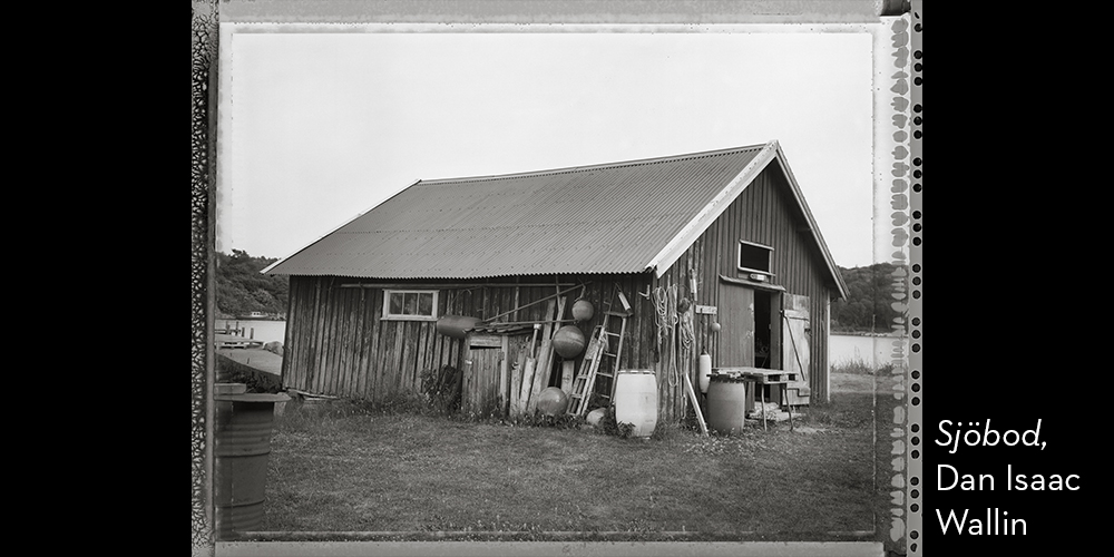 Svartvitt foto. Profilbild av en gammal sjöbod, havet i bakgrunden.