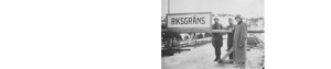 Svartvitt foto. Tre män som står bredvid varandra nära en skylt med text: Riksgräns.