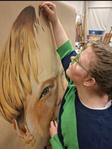 Foto. Person i profil som står till höger sida av bilden och målar på en stor canvas. 